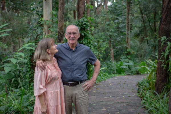 Husband and wife standing on a boardwalk in a forest garden with wife looking up at husband